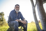 A bearded man sits on a rock outside, next to a tree, with a stern facial expression.