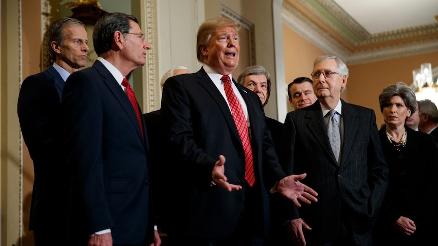 Donald Trump makes an open-handed gesture surrounded by five men and a woman in black suits.