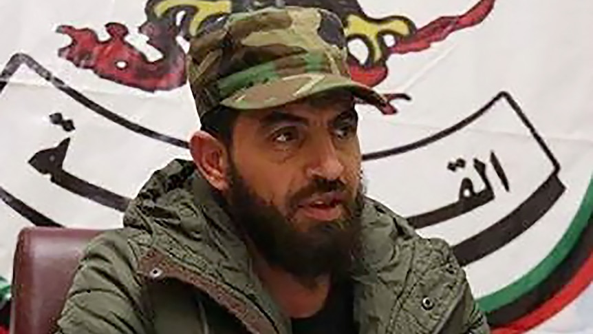 A bearded man in a military cap sits in front of a sign with Arabic script.