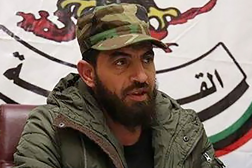 A bearded man in a military cap sits in front of a sign with Arabic script.
