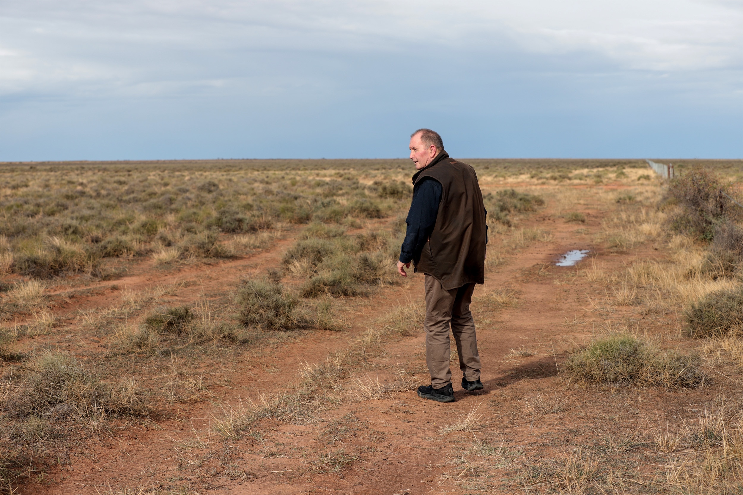 A man wearing a dark coloured shirt and vest stands in a field, looking over his shoulder the sky and horizon visible behind.