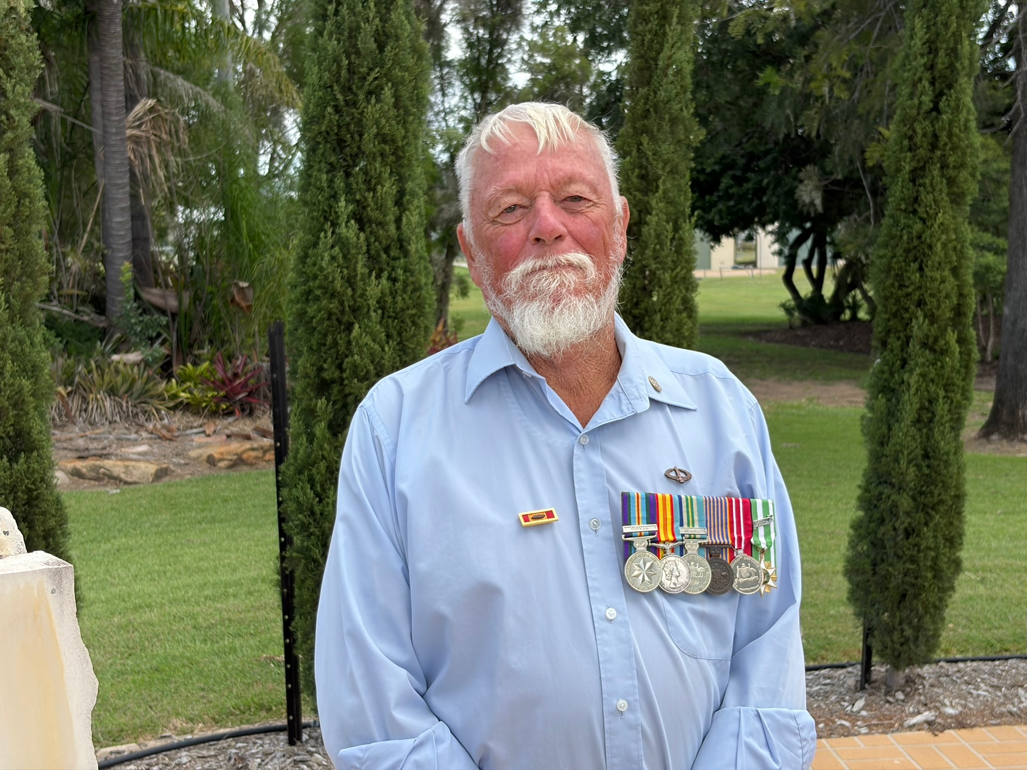 Un hombre de pelo blanco, barba y una hilera de medallas en el pecho se encuentra frente a unos pinos.