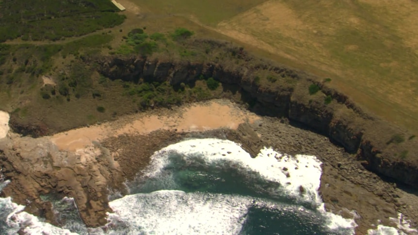 An aerial shot of coastline with greeny-browny grass leading into rugged cliffs above blue and white water.