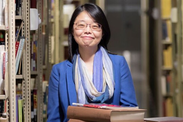 A woman wearing a blue jacket and glasses smiles at the camera inside a library