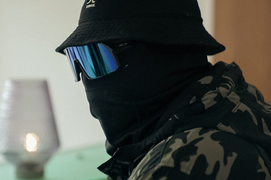 A close up of a man completely covered by a mask over his mouth, sunglasses and a bucket hat.