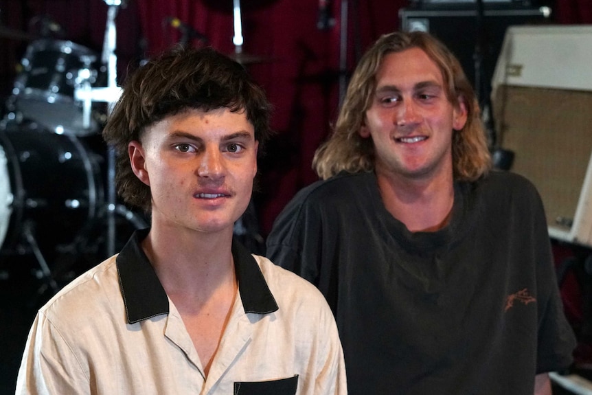 Two men sit in a band room with drums in the background.