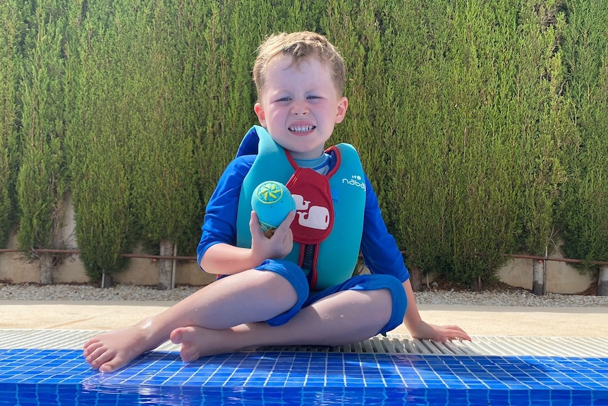 A smiling, fair-haired boy sits poolside in his floaties.