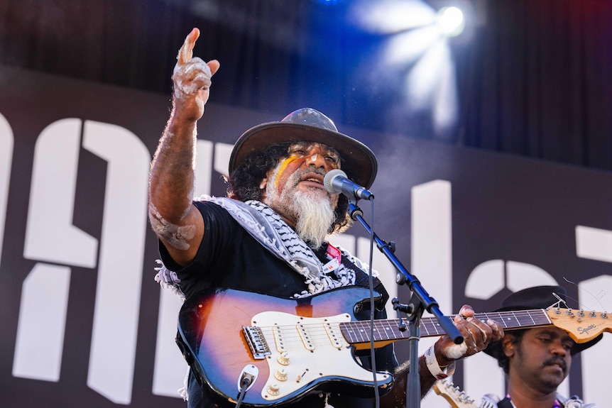 A man with a beard and wide brimmed hat points to the sky with a guitar in hand.