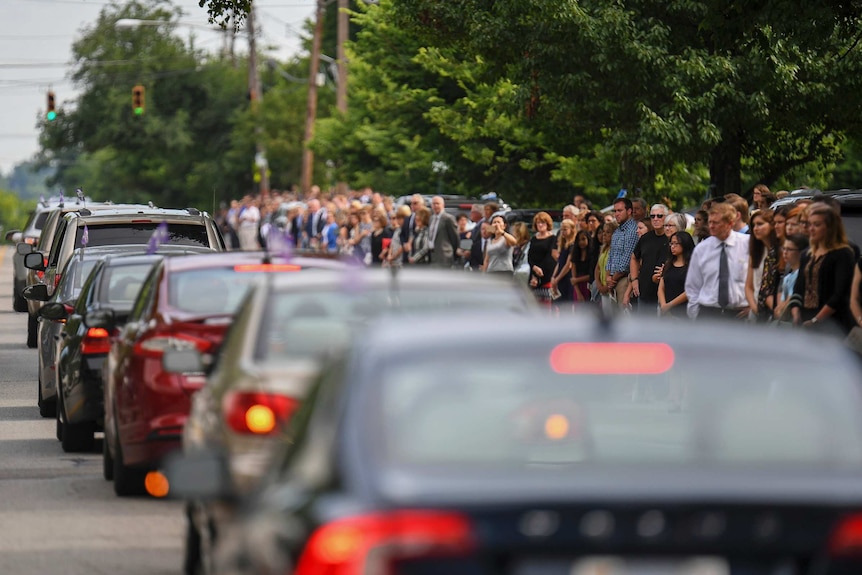 Mourners line the street as a procession of cars drive by.
