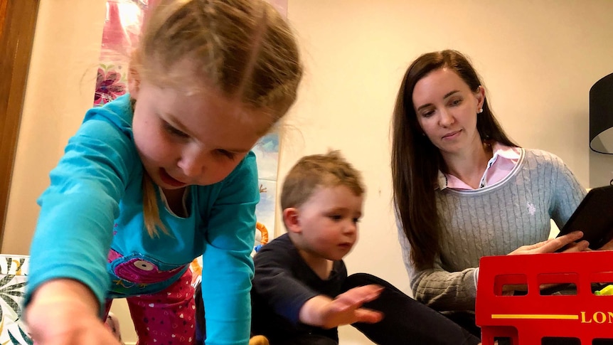 A brunette woman sits on the ground, playing with her two young kids. There are toys on the carpet.