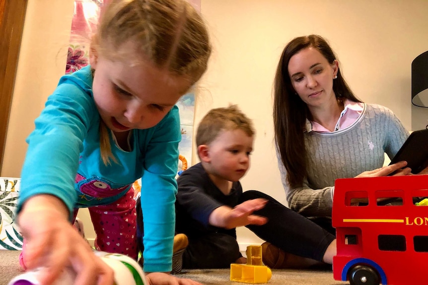 A brunette woman sits on the ground, playing with her two young kids. There are toys on the carpet.