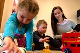 A brunette woman sits on the ground, playing with her two young kids. There are toys on the carpet.