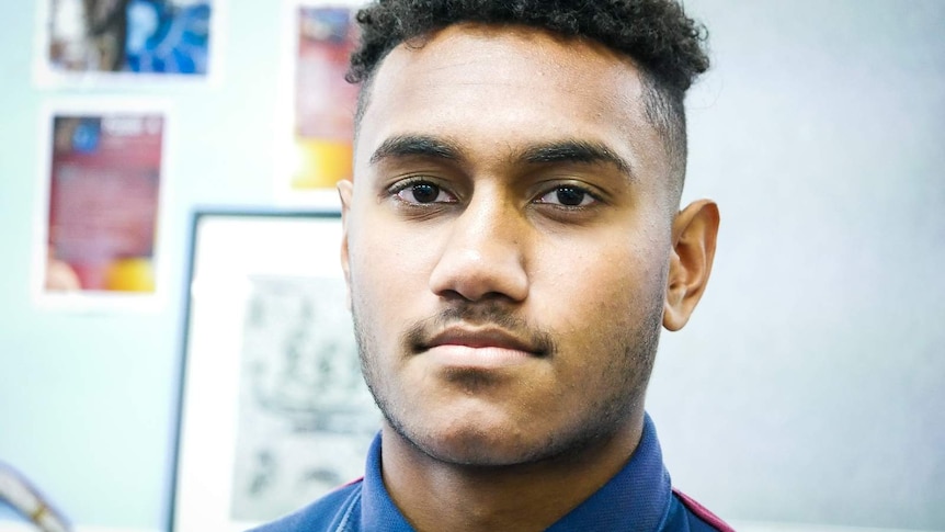 A young man looks right at the camera. There are pictures of South Sea Islanders on the wall behind him.