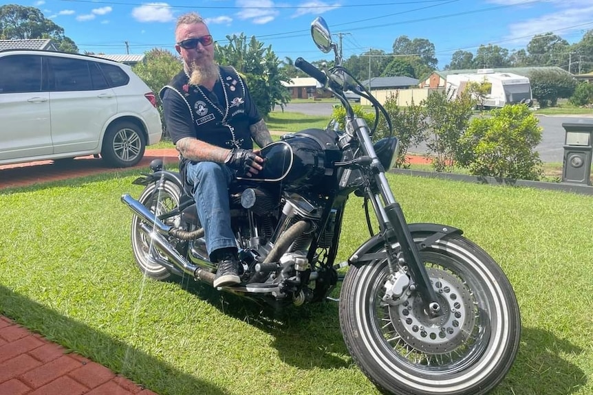 Steve Whiteley with a t-shirt, vest and jeans on, with no helmet, on a motor bike, on a grassy lawn on suburban street 