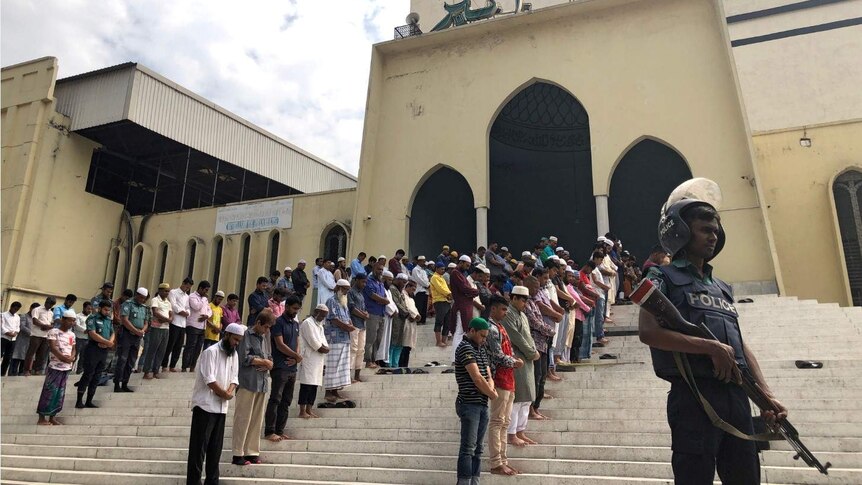 A group of men gathered on a the steps of a mosque, their heads bowed in prayer. A police officer with a gun stands guard.