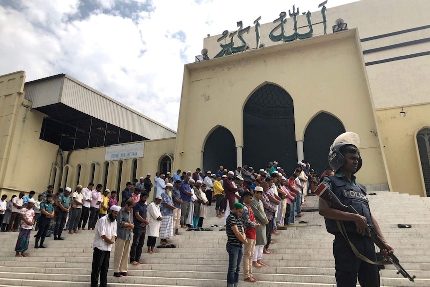 A group of men gathered on a the steps of a mosque, their heads bowed in prayer. A police officer with a gun stands guard.