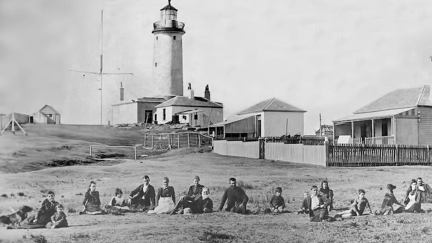 A black and white historical photo of a crowd of people outside a towering lighthouse.