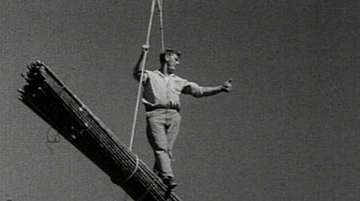 A workman standing on top of building materials suspended by a crane.