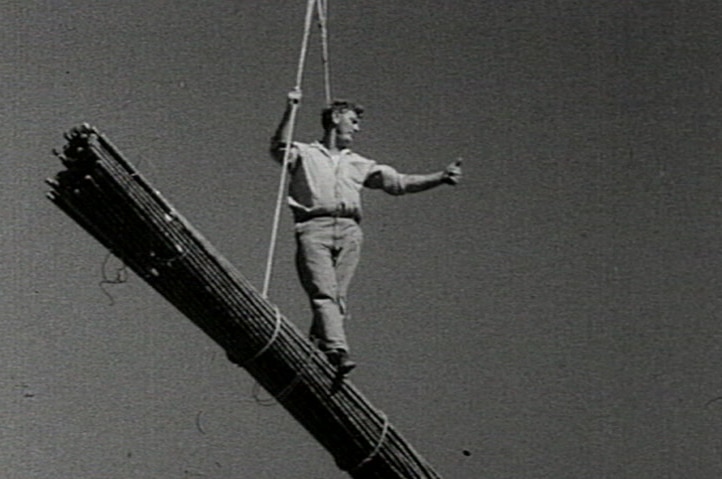 A workman standing on top of building materials suspended by a crane.