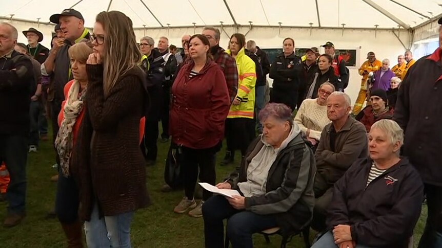 A group of people stands in a tent, many of them wearing emergency services uniforms in bright yellow and orange.