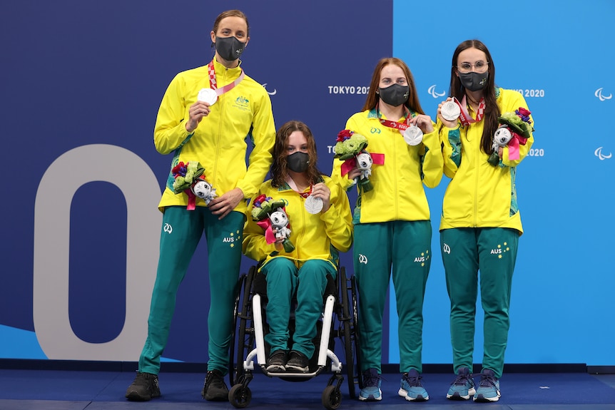 Four Australian swimmers wearing green and gold tracksuits and masks hold their silver medals on the medal dais.