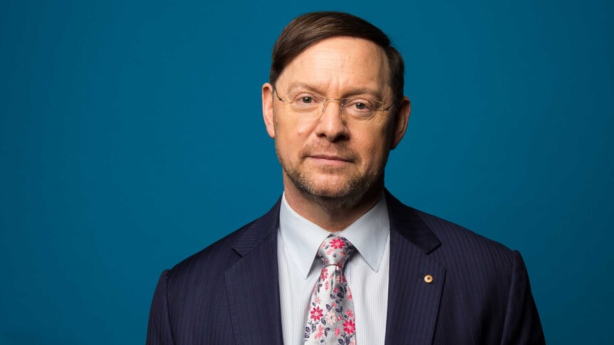 Professor John Rasko AO wearing suit and standing in front of blue background.