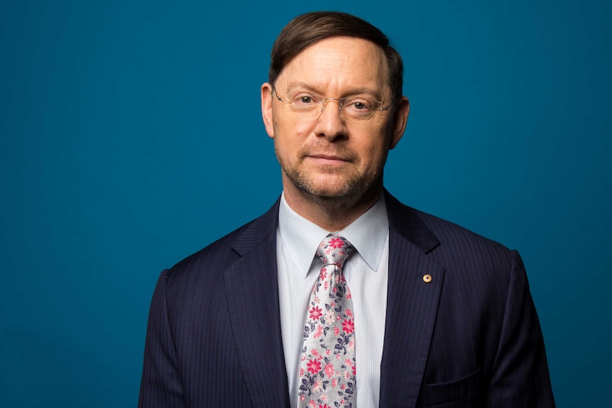 Professor John Rasko AO wearing suit and standing in front of blue background.