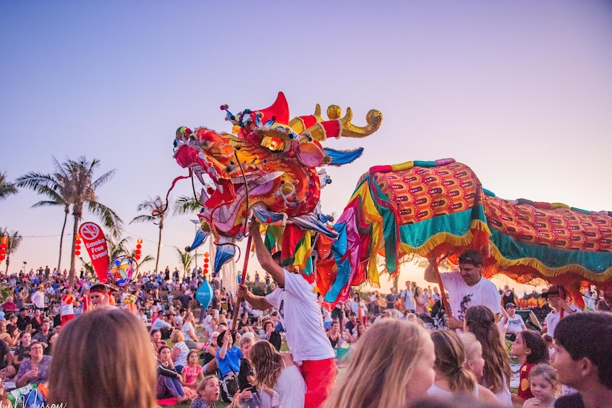 A large Chinese dragon performs for a crowd of people with palm trees in background