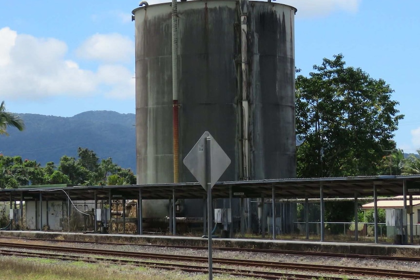 Old molasses tank with rusty pipes and moss and mildew on it, behind a railway line.