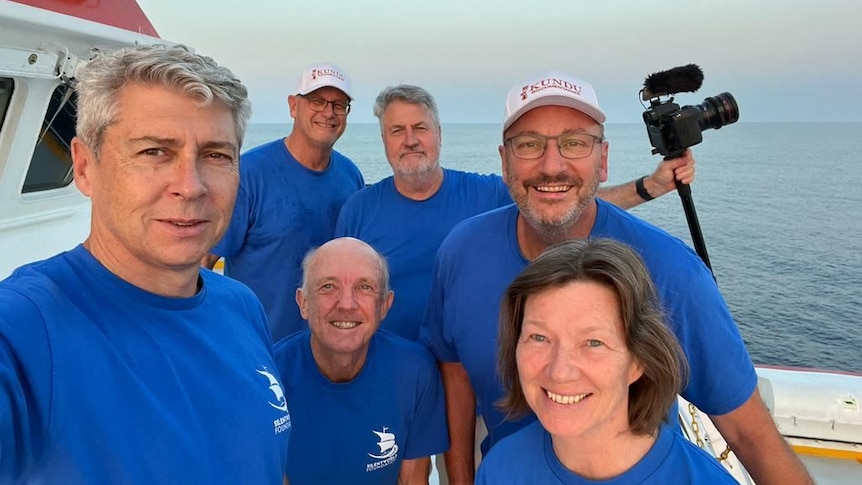A group of people in blue t-shirts smile and pose for a photo while onboard a ship.