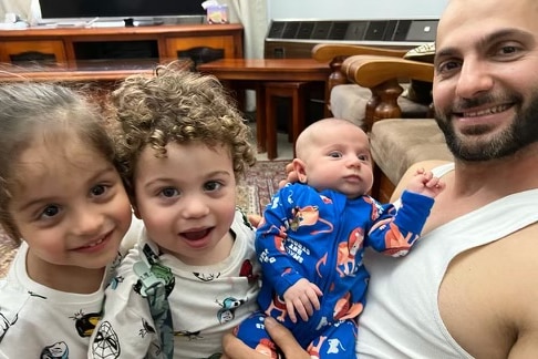 A man holding an infant with a toddler and pre-schooler on his lap. They're all smiling.