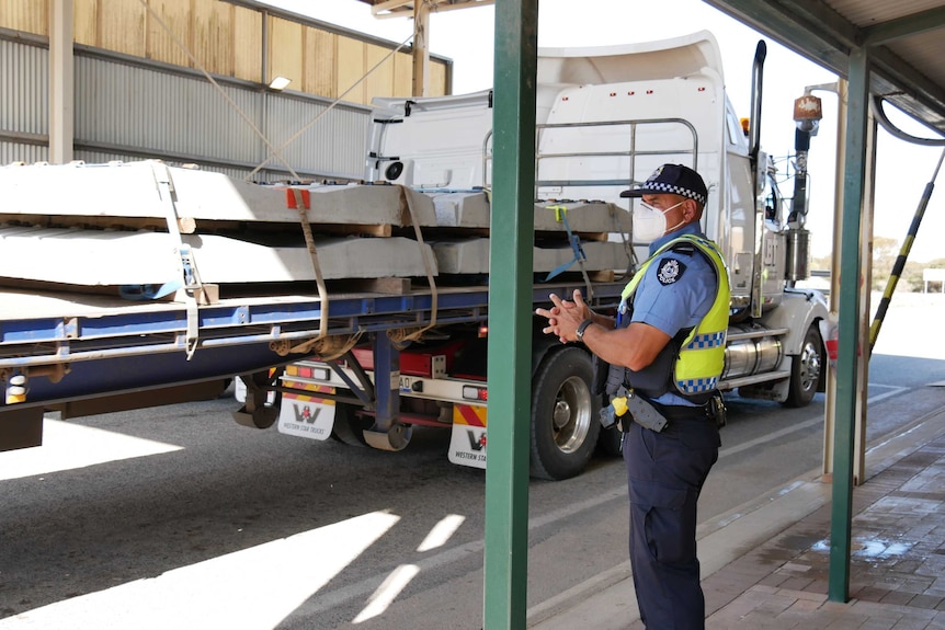 A police officer, wearing a mask, stands under a shed-like structure next to a truck.