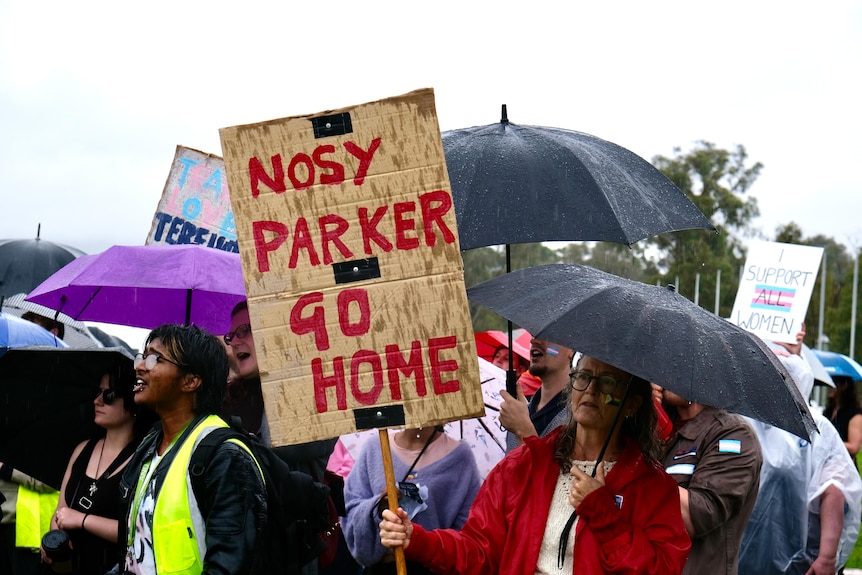 A woman holds a sign saying 'Nosy Parker go home', surrounded by a crowd holding umbrellas in the rain.