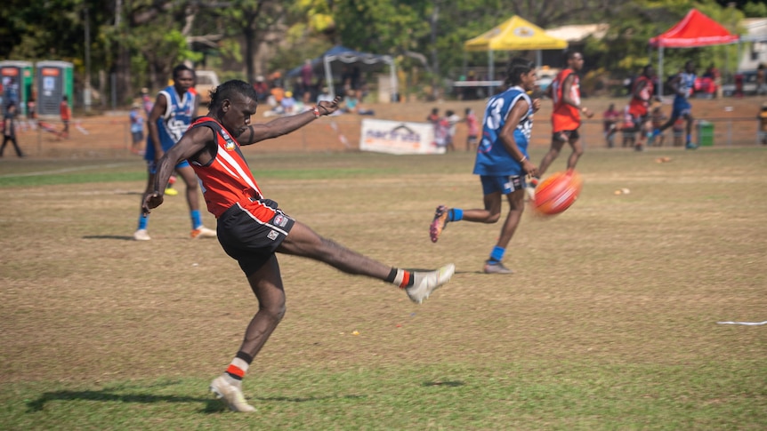 A football player kicking an Aussie rules football on an oval. 