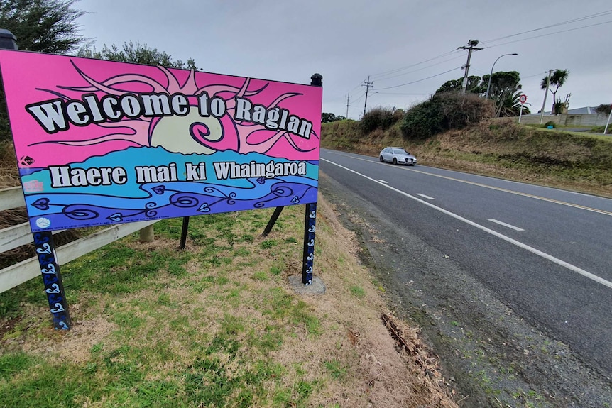 A pink sign next to a road with 'welcome to Raglan' and Maori words written on it.