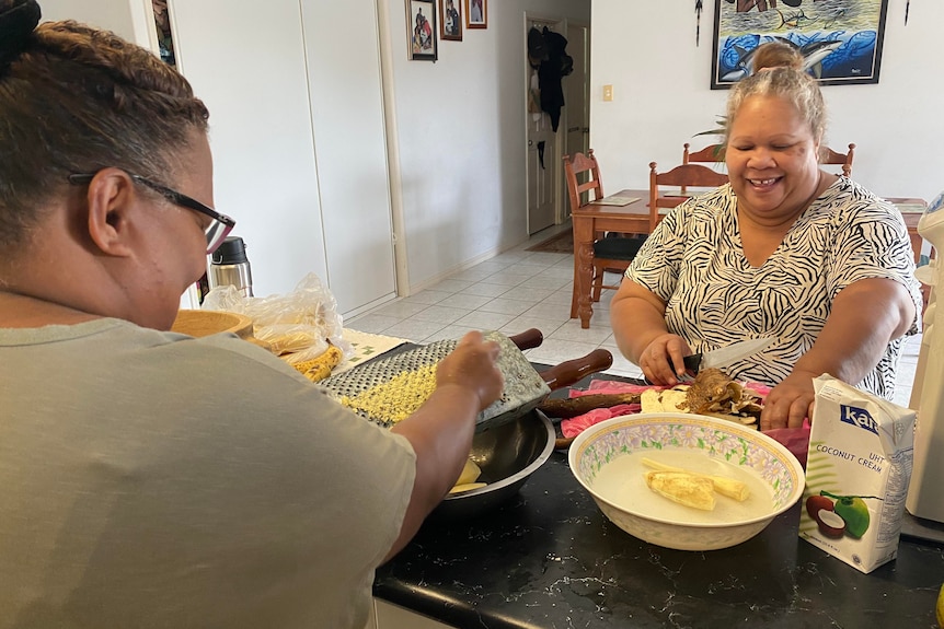 Two women sitting at a kitchen bench mashing bananas in a bowl. 