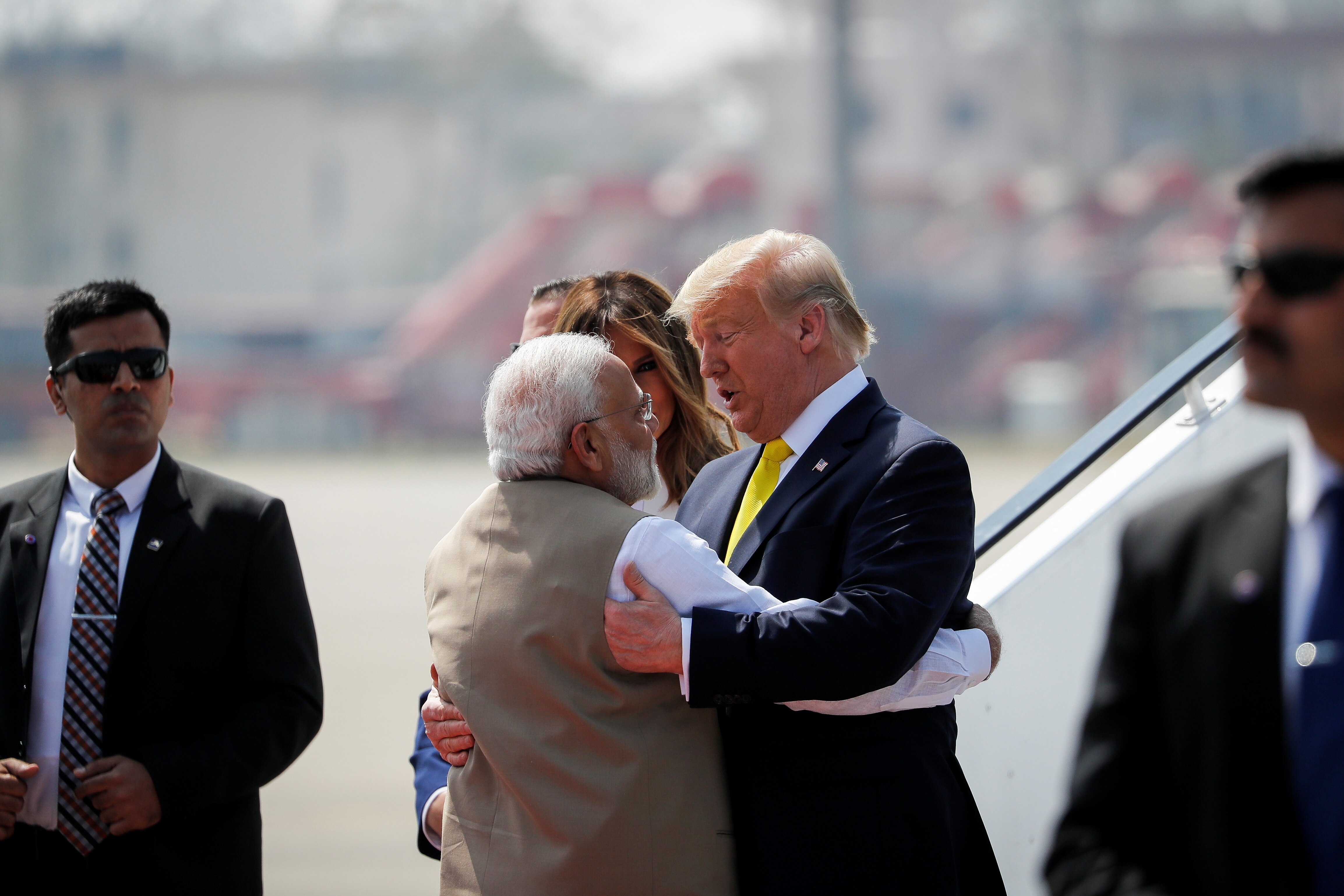 Man with grey hair and beard with back to camera embraces taller man with light hair and orange skin