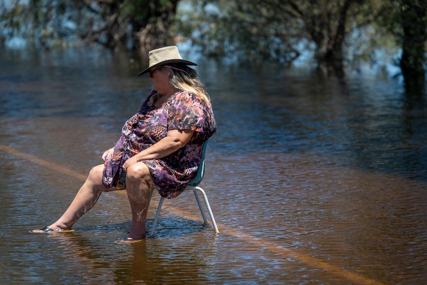 A woman wearing a hat and dress sits on a blue chair on a flooded road, surrounded by water