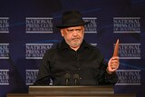 Noel holds up an Indigenous clap stick at the National Press Club speakers podium.