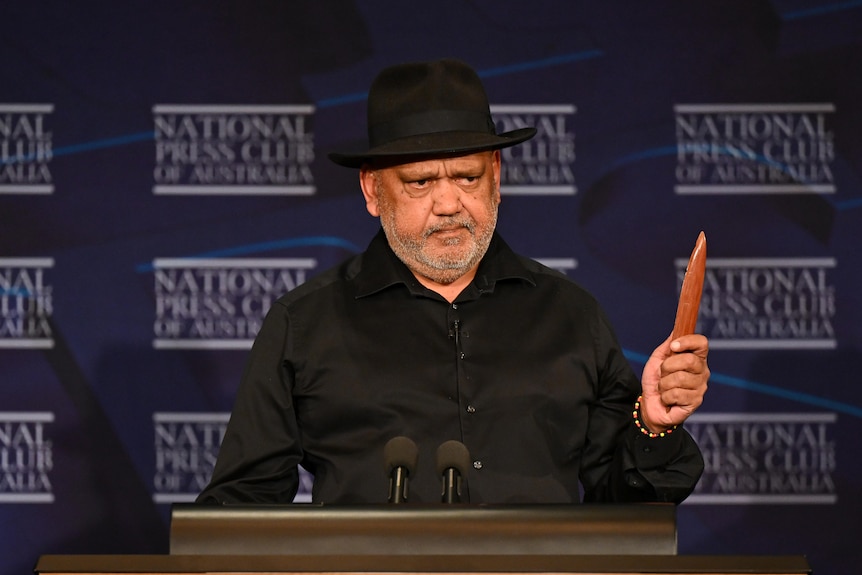 Noel holds up an Indigenous clap stick at the National Press Club speakers podium.