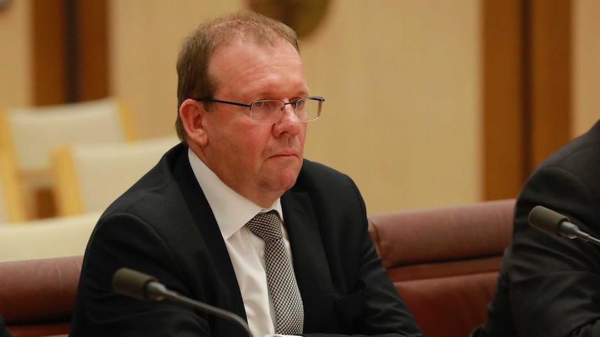 Grant Hehir sits with his arms crossed at a table looking at politicians out of the picture