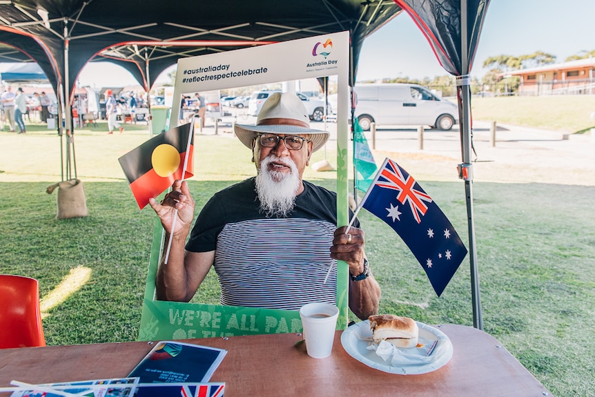 Indigenous man with hat and glasses waves Aboriginal and Australian flags 