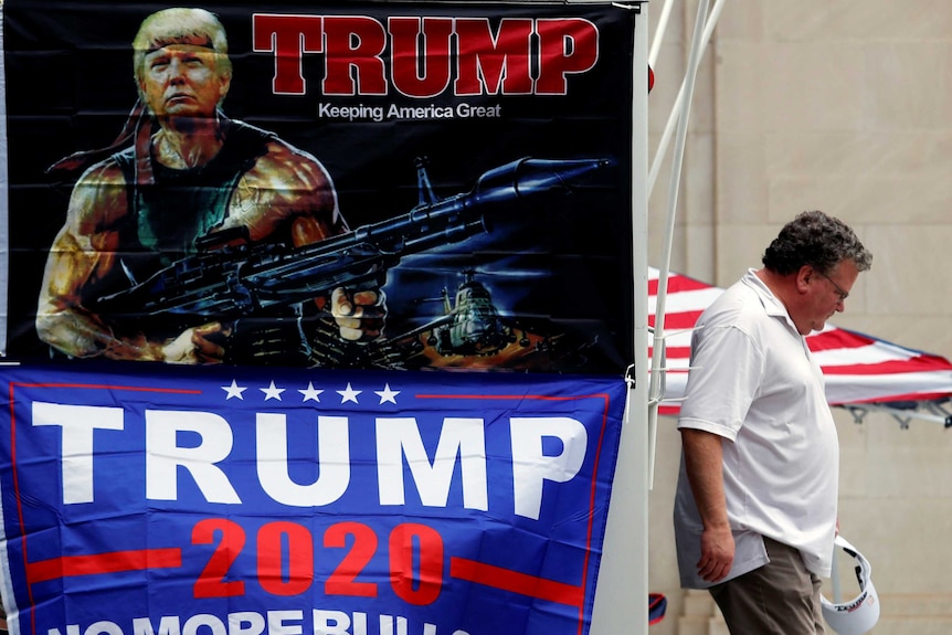 A man carries a white Trump branded visor next to a flag with Donald Trump's face superimposed on the body of Rambo.