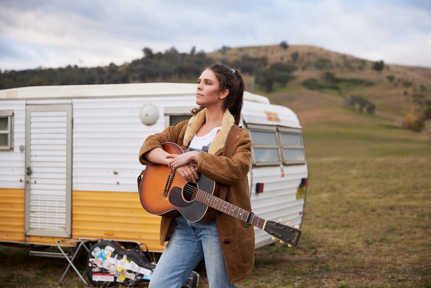 Fanny stands in front of a white and yellow caravan, holding a guitar, with a wide grassy landscape in the background