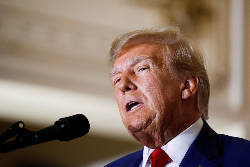 A close up of Trump speaking in a heritage building. He is wearing a dark suit and a red tie over his white shirt.