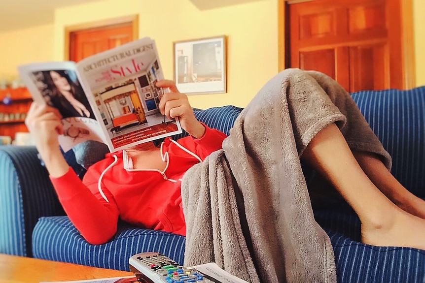 A woman reading a magazine lies on a couch.