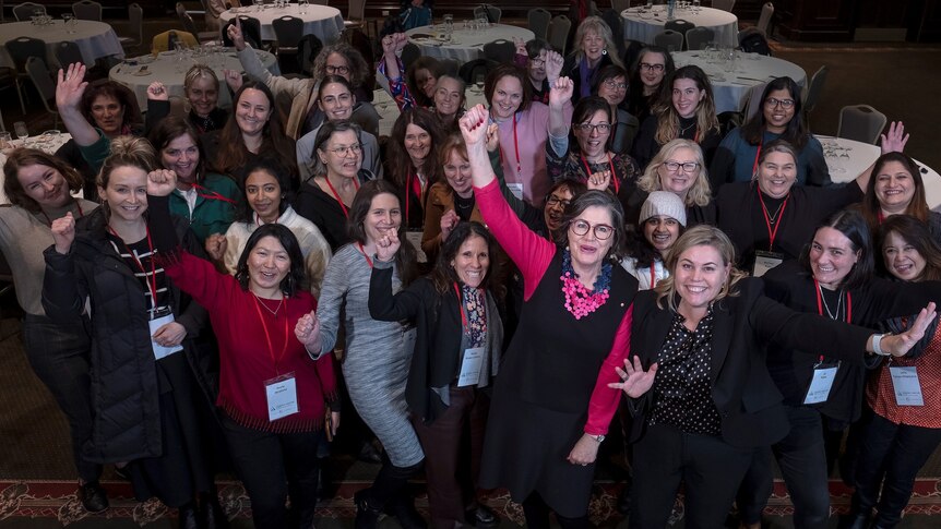 A group of women in a conference room look up at a camera and raise their arms in the air in a celebratory way