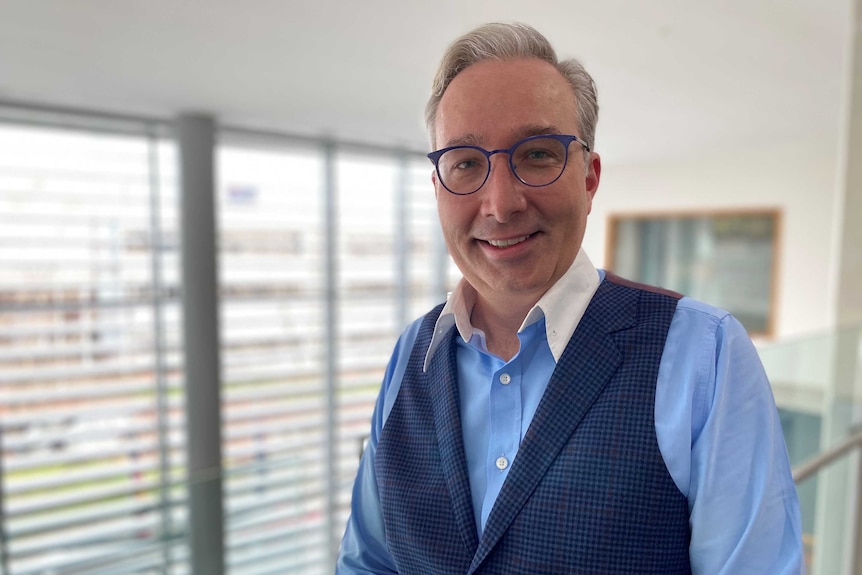 Man in blue shirt, blue waistcoat and blue glasses looks at camera, smiling