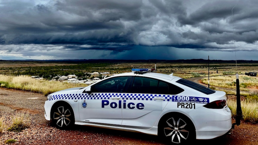 A blue and white WA police car parked on red dirt overlooking buildings under ominous grey storm clouds.