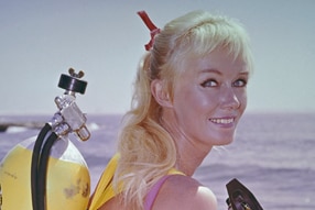Archival photo of Valerie Taylor at beach with blonde ponytail and in pink swimsuit wearing yellow scuba tank.
